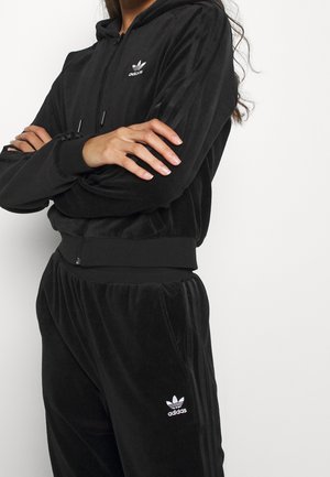 Persoon draagt een zwart Adidas velours trainingspak met logo op borst en dij, armen gekruist tegen een witte achtergrond.