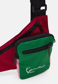 Kolorowa torebka na ramię, z czerwoną górną częścią i zieloną dolną komorą z różowymi akcentami. Zawiera dwie kieszenie na zamek.