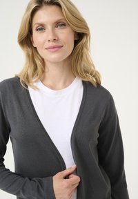 Šedý cardigan vyrobený z měkké tkaniny, s hlubokým výstřihem do V a dlouhými rukávy, přeložený přes obyčejné bílé tričko, ruce držící cardigan.