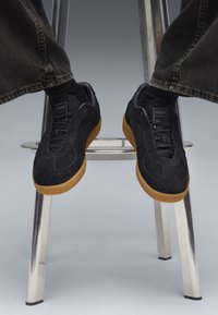 Svarta mockaskor med låg profil och gummisula, med snörningsdesign och texturerade detaljer. Bärs med mörka jeans.