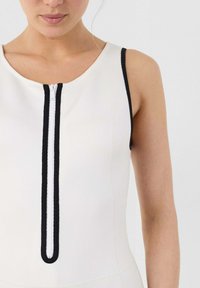 Prenda blanca sin mangas con cremallera frontal, ribete negro contrastante en el escote y la cremallera, textura suave, diseño ajustado.