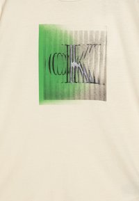 Krämfärgad t-shirt med ett tryckt mönster i grön och grå gradient, som visar abstrakta former och linjer i varierande tjocklekar.