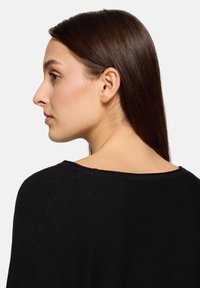 Femme aux cheveux bruns raides portant un pull côtelé noir, vue de profil et de dos sur un fond uni clair.