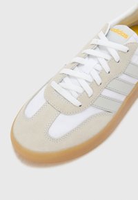 Adidas tenisice s bijelim kožnim gornjim dijelom, bež zamščenim detaljima, sivim prugama i gumenim potplatom. Ima bijele vezice i detalj s logom.
