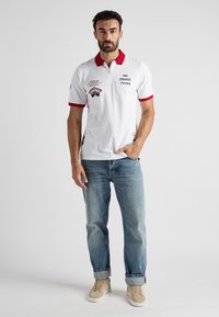 Biała koszulka polo z czerwonym kołnierzykiem i mankietami, z haftem na piersi i małą kieszonką. Nosi ją w połączeniu z jasnoniebieskimi dżinsami i beżowymi sneakersami.