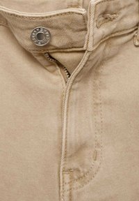 Gros plan sur un pantalon beige avec une fermeture éclair partiellement ouverte et un bouton en métal fixé à la taille.