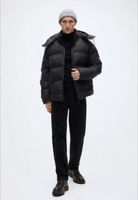 Geacă matlasată neagră cu fermoar frontal și glugă, asortată cu un pulover negru cu guler înalt, pantaloni negri și căști de piele maro. Căciulă gri deschis.