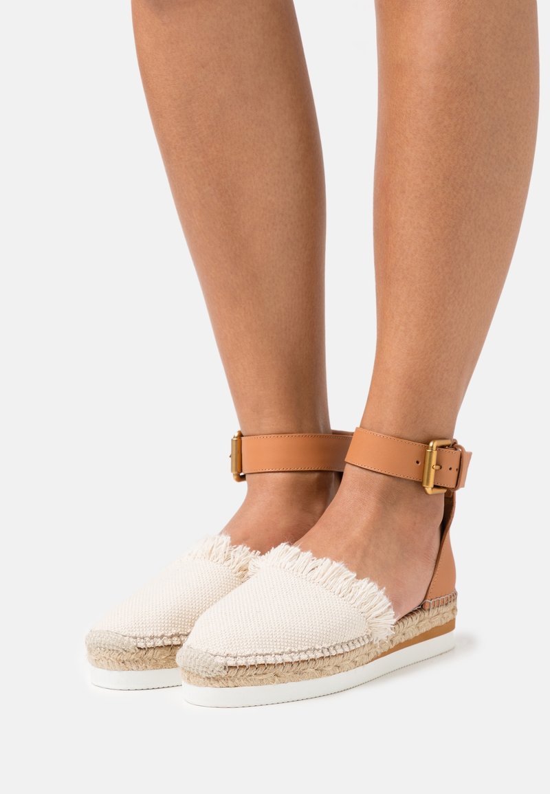 Pieds portant des espadrilles plates beiges avec des détails à franges et des brides de cheville couleur cognac avec boucles dorées sur fond blanc.