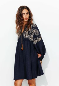 Robe bleu marine à coupe décontractée, ornée de broderies florales et de franges au niveau du col. Manches longues et amples, en tissu texturé.