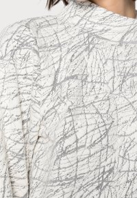 Valkoinen kangas, jossa mustia, abstrakteja viivoja muistuttava teksturoitu kuvio. Mallissa on korkea kaulus ja pehmeä, kevyt materiaali.