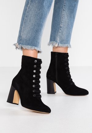 Schwarze Wildleder-Stiefeletten mit dekorativen Knöpfen und Blockabsätzen, getragen zu hellblauen Jeans mit ausgefranstem Saum auf weißem Boden.