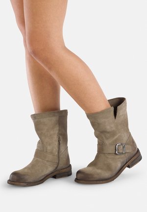 Personne portant des bottes en cuir marron usées mi-mollet avec des détails de fermeture éclair et de boucle, croisant une jambe sur l'autre.