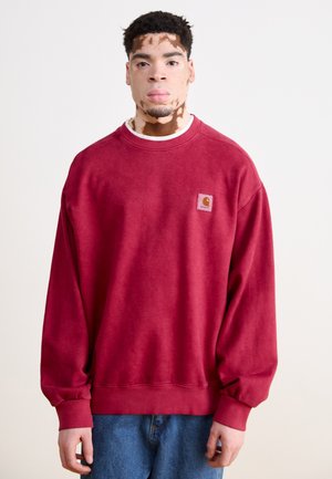 Sudadera carmesí de manga larga, con puños y dobladillo acanalados; presenta un pequeño parche de marca en el pecho; usada sobre una camisa blanca.