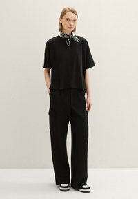 Schwarzes Kurzarmshirt mit geradem Schnitt, kombiniert mit weitgeschnittenen schwarzen Hosen mit Seitentaschen und schwarz-weißen Sneakers.