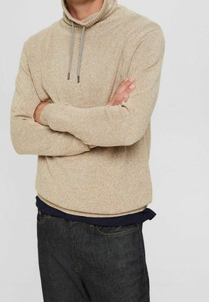Maglione a maglia beige con colletto alto, dotato di un cordino di colore a contrasto. Indossato sopra una camicia blu navy, abbinato a jeans in denim scuro.