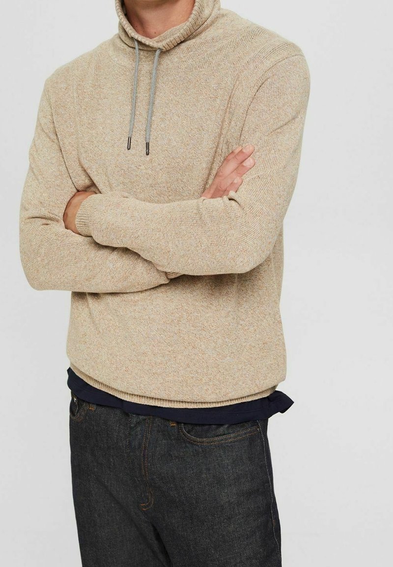 Pull en tricot beige avec un col montant, doté d'un cordon de serrage de couleur contrastante. Porté par-dessus une chemise marine, associé à un jean en denim foncé.