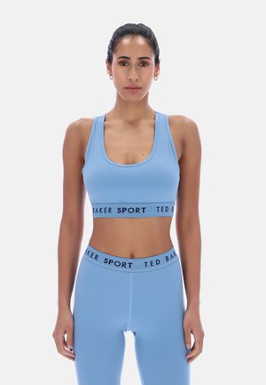Kvinne stående vendt framover iført lys blå sports-BH og leggings, begge med "TED BAKER SPORT" merke på elastiske bånd.