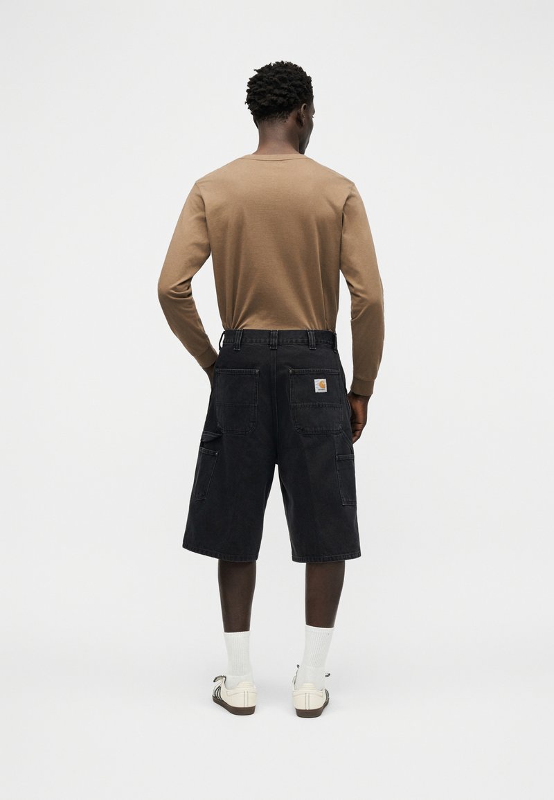 Uomo in piedi di spalle che indossa una camicia a maniche lunghe color sabbia, pantaloncini cargo neri fino al ginocchio, calzini bianchi e sneakers bianche basse.