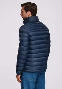 Marineblaue Steppjacke mit horizontalem Quiltmuster, hohem Kragen und elastischen Bündchen, kombiniert mit blauen Jeans. Rückansicht gezeigt.
