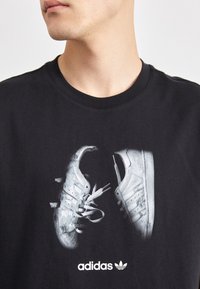 T-shirt noir avec un graphique de deux baskets blanches avec des lacets. Le design comprend le logo Adidas en blanc en bas.