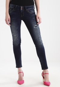 Jeans ajustados de color azul oscuro con detalles rasgados, que cuentan con un cierre de dos botones y costuras en contraste. Combinados con unos zapatos de tacón alto rosa.