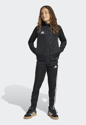 Chándal atlético negro con rayas blancas, que incluye una chaqueta con cremallera y pantalones ajustados, combinado con zapatillas negras y una tela texturizada.