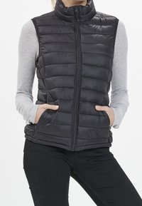 Černá nadýchaná vesta s vysokým límcem, zapínáním na zip a dvěma předními kapsami. Povrch zdobí prošité panely pro texturu. Vhodná na vrstvení.
