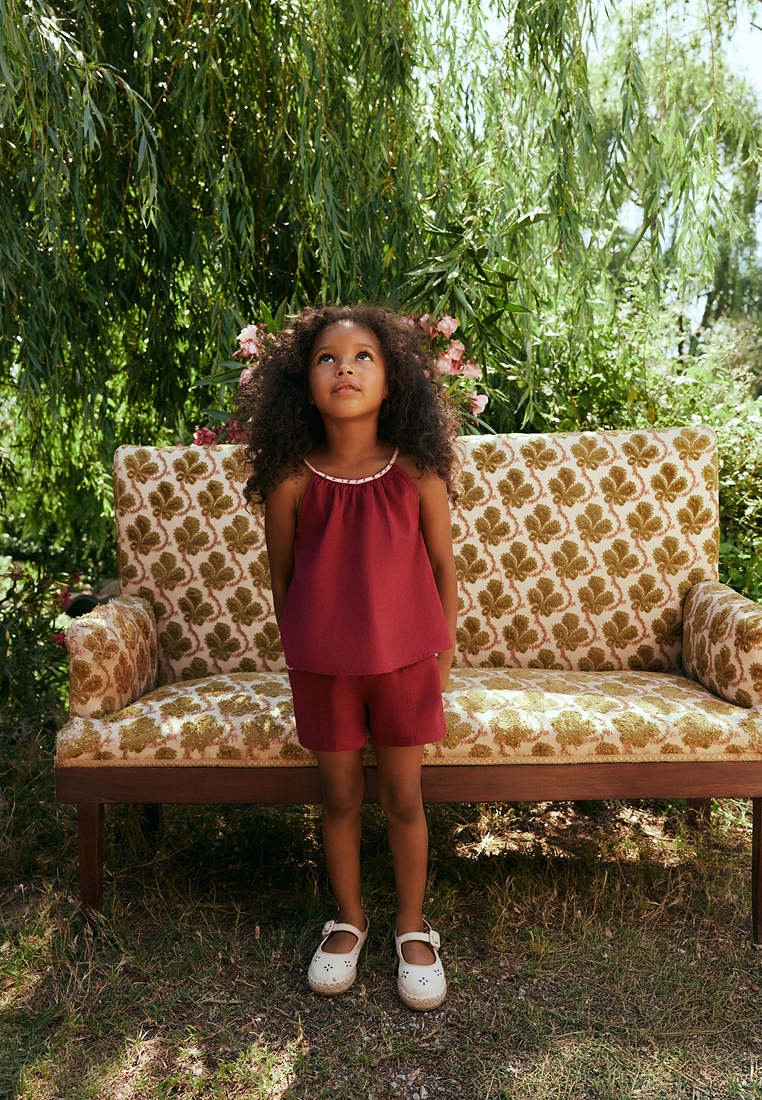 Jeune fille aux cheveux bouclés vêtue de rouge, debout devant un banc à motif floral en plein air sous des arbres verts.