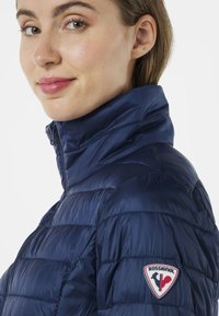 Mörkblå isolerad jacka med hög krage, tuffat mönster och en framträdande Rossignol-logotyp på vänster axel.