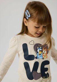 Lys beige langermet skjorte med "LOVE" grafikk i mørkeblå, med tegneseriehunder. Har detaljer med puff ermer og gullakenter.