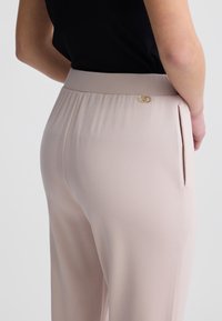Beige Hosen mit glatter Textur, ausgestattet mit einem goldenen Akzent nahe dem Bund und seitlichen Taschen. Taillierte Passform, leicht lockerer Schnitt.