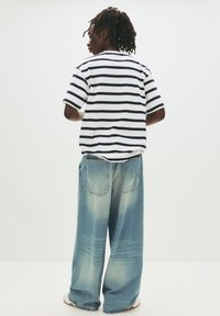 Persoon met dreadlocks, gekleed in een wit en zwart gestreept T-shirt en losse vervaagde blauwe jeans, staand tegen een effen witte achtergrond.