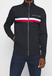 Černá zipová bunda s vysokým límcem, zdobená horizontálními pruhy v červené, bílé a námořnické modré na hrudi. Měkký, lehký materiál.