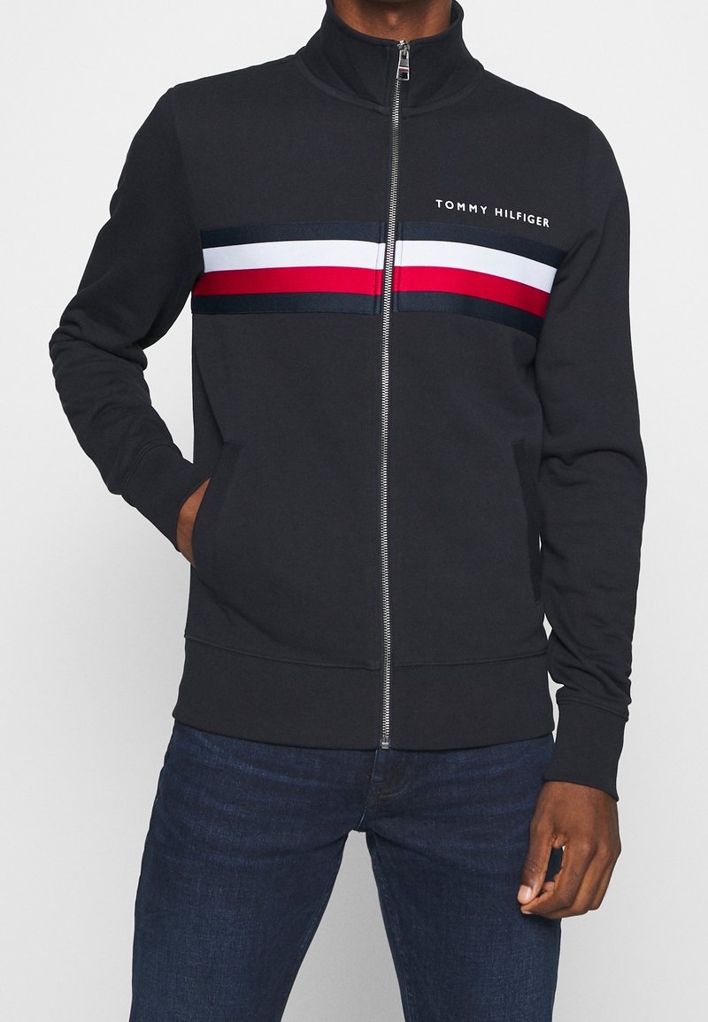 Černá zipová bunda s vysokým límcem, zdobená horizontálními pruhy v červené, bílé a námořnické modré na hrudi. Měkký, lehký materiál.