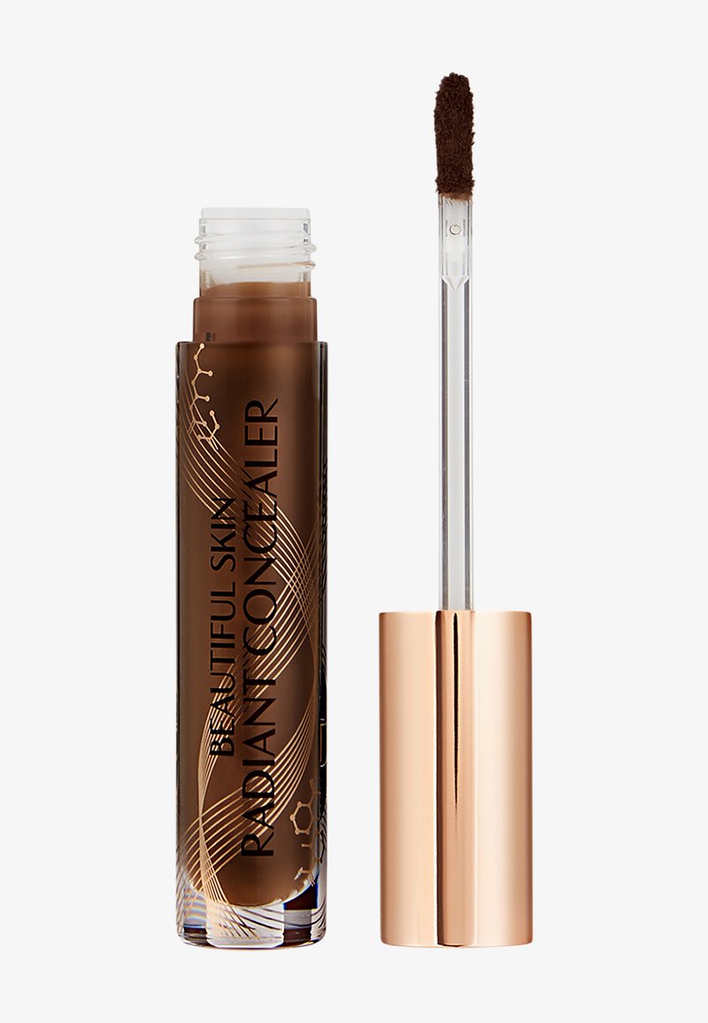 Tube de correcteur avec un produit liquide marron et un applicateur transparent muni d'une éponge en forme de patte de biche, à côté d'un bouchon en or rose.