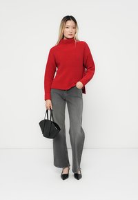 Roter geraffter Strickpullover mit hohem Kragen, kombiniert mit grauen Schlagjeans. Das Modell hält eine schwarze Tragetasche und trägt spitze schwarze Schuhe.