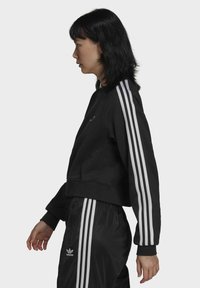 Svart kort sweatshirt med vita tre ränder på varje ärm. Bärs med matchande svarta träningsbyxor med en liten logotyp och ficka med dragkedja.