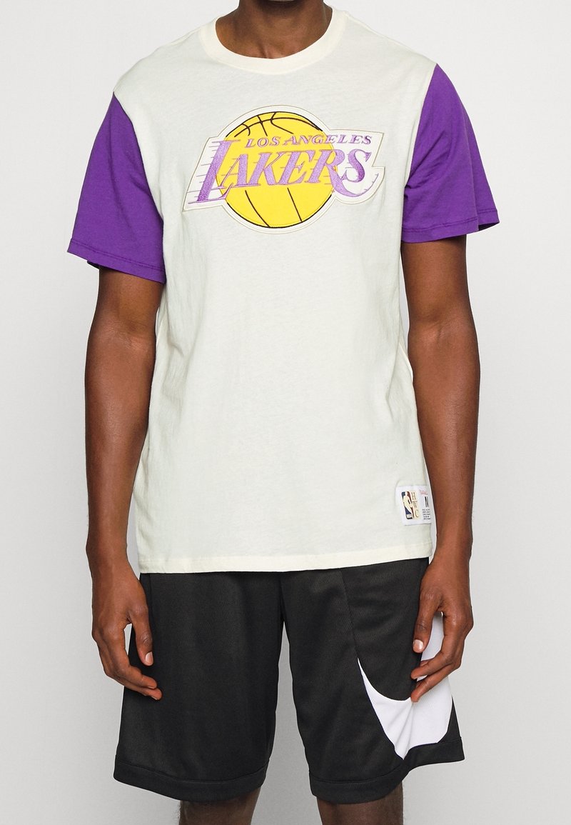 Camiseta de manga corta con tejido crema y morado, con un gran logo de "Los Angeles Lakers" y un gráfico de baloncesto en la parte frontal.