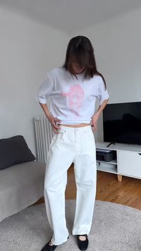 Γυναίκα προσαρμόζει το λευκό της παντελόνι, φοράει ένα λευκό μπλουζάκι με ροζ λεοπάρ μοτίβο, στέκεται σε μοντέρνο σαλόνι με τηλεόραση και καναπέ.