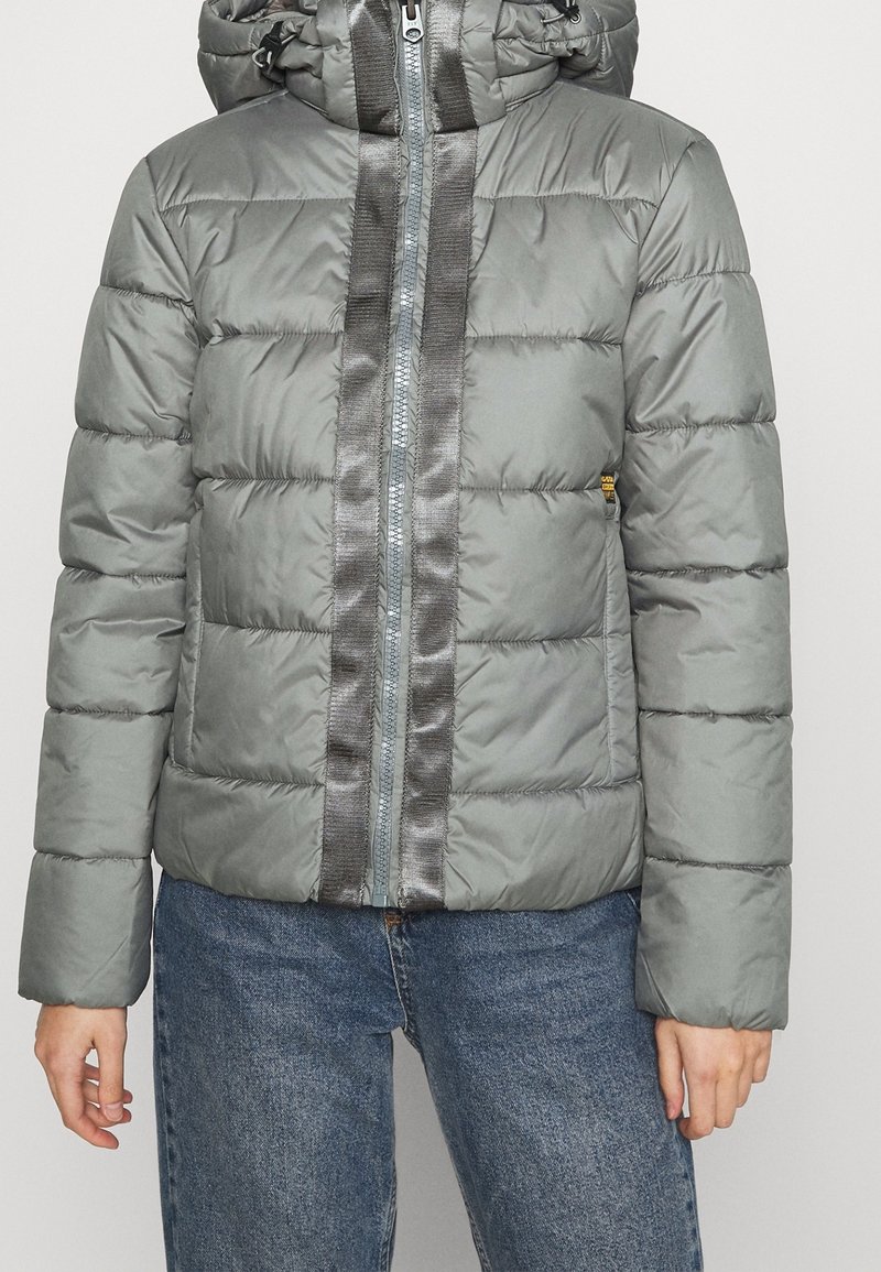 Šedá péřová bunda s lesklým předním pruhem, kapucí a elastickými manžetami, obsahující zipový uzávěr a prošívanou strukturu.