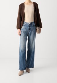 Brauner, flauschiger Cardigan, beigefarbenes, eng anliegendes Oberteil, weit geschnittene, hellblaue Jeans und nudefarbene, spitz zulaufende Absätze. Kleine Tasche in der Hand.