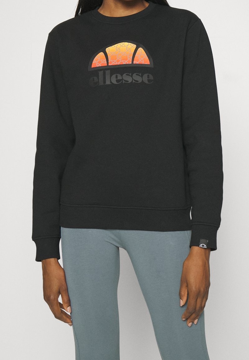 Personne portant un sweat-shirt noir ellesse avec un logo demi-cercle orange et des leggings bleu clair, debout devant un fond uni.