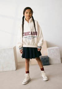 Krém fleece pulóver, amelyen piros hímzés szerepel: "CHICAGO Lake Michigan". Navy kék pliszírozott szoknyával és fehér sportcipővel, piros zoknikkal párosítva.