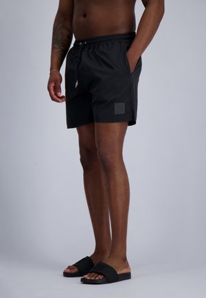 Man die zwarte shorts met trekkoord en zwarte slides draagt, staat met één hand in zijn zak tegen een effen achtergrond.