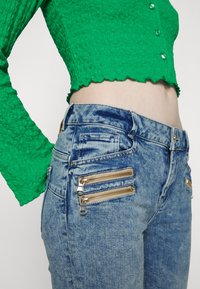 Top de manga larga texturizado verde combinado con unos jeans de mezclilla azul que presentan tres cremalleras decorativas doradas en los bolsillos laterales.