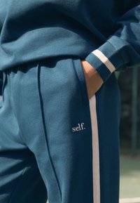 Türkisfarbene Jogginghose mit weißen Seitenstreifen. Das Material wirkt weich und strukturiert. Auf dem Taschenbereich befindet sich ein kleines, gesticktes "self."-Logo.