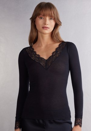 Kvinde iført en tætsiddende sort langærmet top med blonder på V-udskæringen og ærmekanterne, stående foran en ensartet lys baggrund.