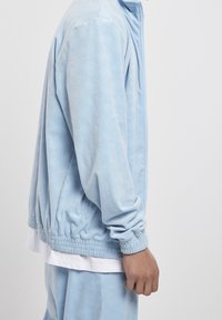 Světlomodrá bunda na zip s texturovaným vzorem, s elastickými manžetami a pasem. Kombinováno s bílým tričkem pod ní.