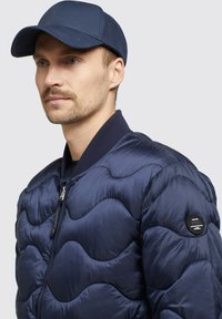 Marinblå puffjacka med vågig quiltad design, dragkedjestängning och ribbad krage. Matchas med en marinblå keps.