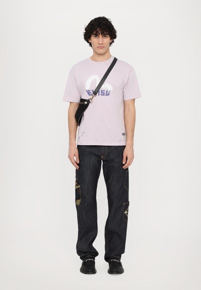 T-shirt en coton lilas clair avec impression graphique, associé à un jean en denim foncé avec des patchs texturés et des accents dorés. Sac bandoulière noir.